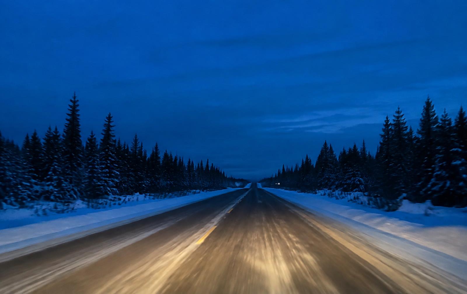 Snow-lined northern highway at dusk with headlights on the road